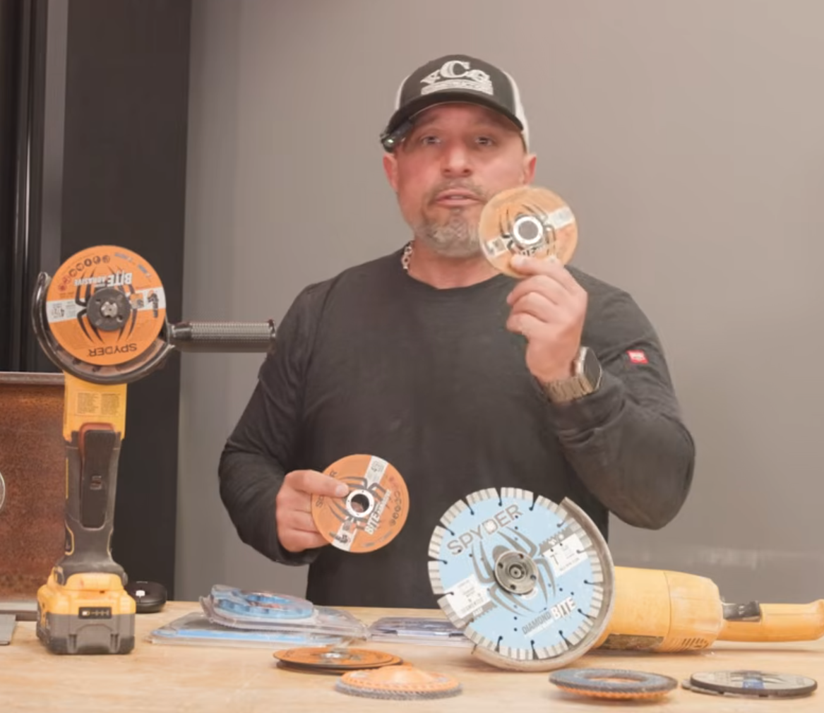 A man wearing a black long-sleeve shirt and a black-and-white baseball cap holds two circular Spyder brand grinding discs. On the table in front of him are several other cutting and grinding discs, a yellow and black cordless grinder, and a yellow large blade cutting tool.